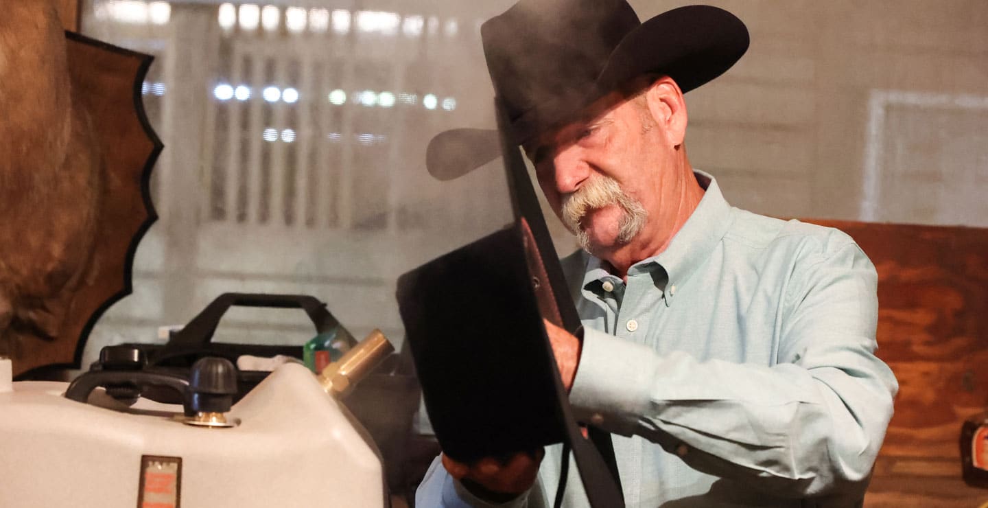 A cowboy with a gray mustache holding an unshaped black hat and placing it on the steamer.